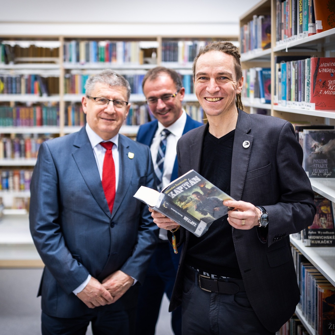 Ivan Bartoš na prohlídce nové knihovny v Roudnici nad Labem. Drží knihu a usmívá se. S ním na fotografii zástupci Roudnice.